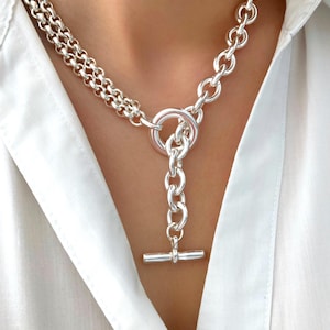 May include: A silver chain necklace with a large, chunky chain and a smaller, delicate chain. The necklace has a toggle clasp and a large, round ring in the center.