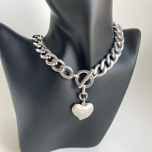 K&ouml;nnte beinhalten: Eine silberne Kettenkette mit einem gro&szlig;en, klobigen Gliederdesign. Die Kette hat einen Toggle-Verschluss und einen baumelnden Herz-Anh&auml;nger.