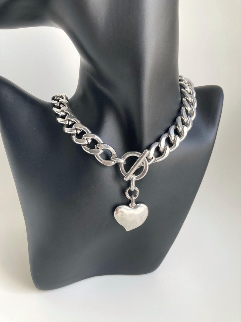 K&ouml;nnte beinhalten: Eine silberne Kettenkette mit einem gro&szlig;en, klobigen Gliederdesign. Die Kette hat einen Toggle-Verschluss und einen baumelnden Herz-Anh&auml;nger.