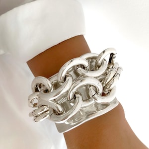 Peut inclure: Gros bracelet argenté avec un design de maillons de chaîne audacieux. Le bracelet est porté au poignet, avec une manche de chemise blanche en arrière-plan. Les maillons sont larges et reliés, créant un bijou de caractère.