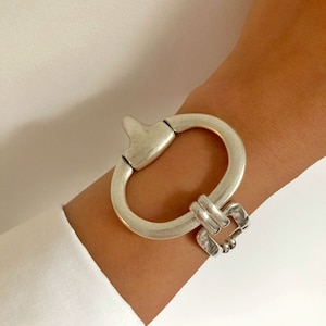 Peut inclure: Un bracelet argenté avec un grand maillon de forme ovale et un fermoir carré plus petit. Le bracelet est en métal et a une finition polie.