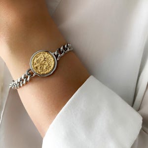 Könnte beinhalten: Ein silbernes Kettenarmband mit einem goldenen Münzanhänger. Die Münze hat ein detailliertes Design und ist in einen silbernen Rahmen gefasst. Das Armband wird am Handgelenk getragen, vor einem weißen Hemd.
