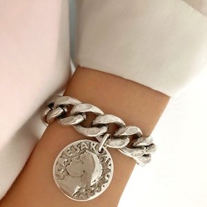 Könnte beinhalten: Silbernes Kettenarmband mit einem großen silbernen Münzenanhänger, der ein Profil von Caesar zeigt. Die Münze trägt die Inschrift "CESAR AUGUSTUS".