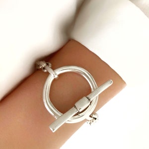 Op de afbeelding: Zilveren armband met een grote, ronde toggle-sluiting. De sluiting bestaat uit twee in elkaar grijpende cirkels, waarvan er één een staaf heeft die door de andere glijdt.