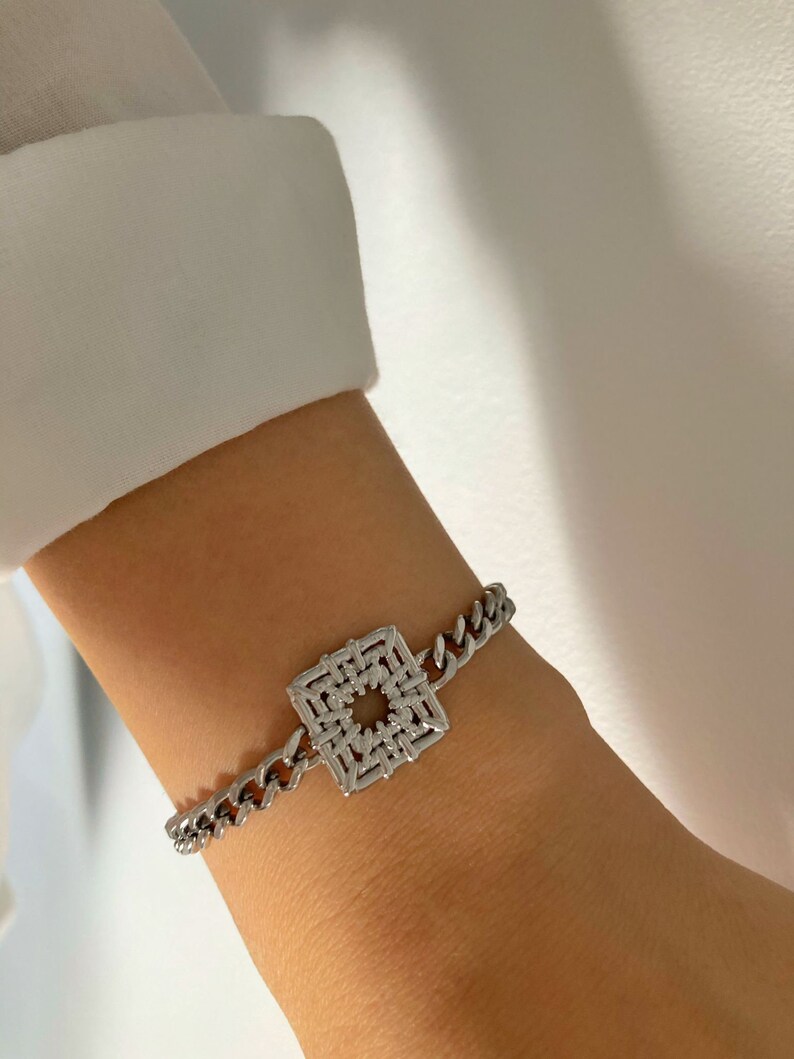 Puede incluir: Un brazalete de cadena de plata con un dije cuadrado con un dise&ntilde;o geom&eacute;trico.