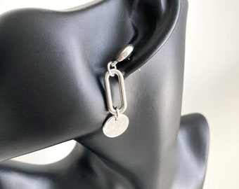 May include: Silver chain earrings with a round charm. The earrings are on a black mannequin head.
