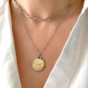 May include: A layered necklace featuring a silver chain with a gold pendant. The pendant is a gold coin design with a textured surface and intricate details. The necklace is worn against a white shirt.