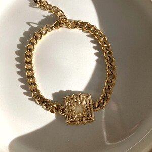 Puede incluir: Un brazalete de cadena dorada con un colgante cuadrado y geom&eacute;trico. El colgante tiene un dise&ntilde;o tejido y una peque&ntilde;a piedra blanca en el centro.