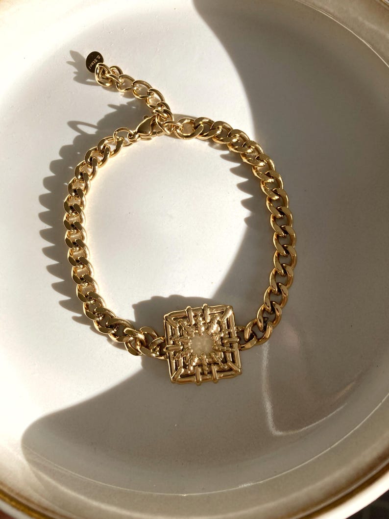 Puede incluir: Un brazalete de cadena dorada con un colgante cuadrado y geom&eacute;trico. El colgante tiene un dise&ntilde;o tejido y una peque&ntilde;a piedra blanca en el centro.