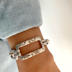 May include: A silver bracelet featuring a large, rectangular, hammered link. The bracelet is made of a chain with multiple smaller links.