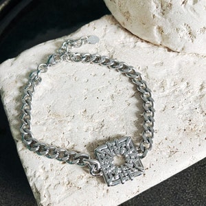 Puede incluir: Un brazalete de cadena de plata con un colgante de forma cuadrada. El colgante est&aacute; hecho de un dise&ntilde;o de metal tejido con un agujero en el centro.