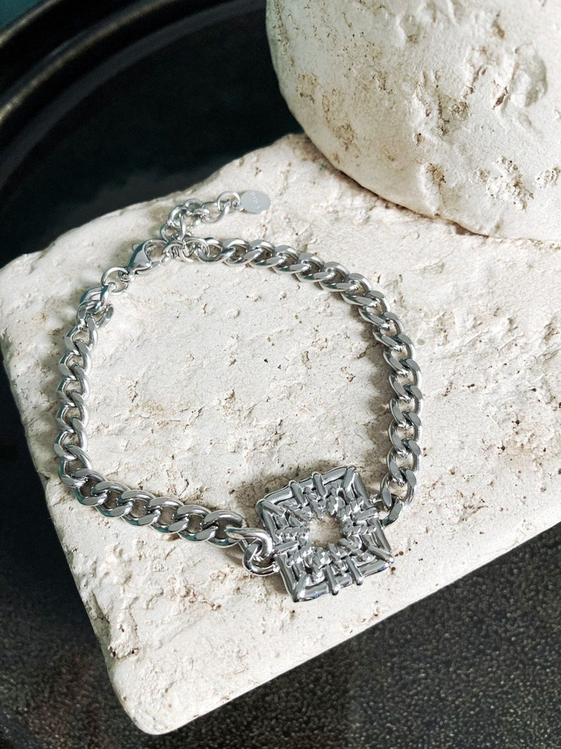Puede incluir: Un brazalete de cadena de plata con un colgante de forma cuadrada. El colgante est&aacute; hecho de un dise&ntilde;o de metal tejido con un agujero en el centro.