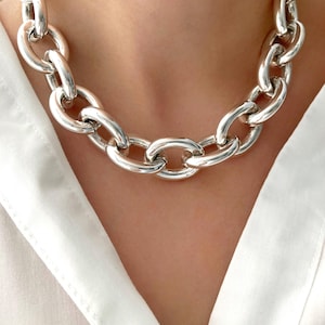 May include: A chunky silver-toned chain necklace with large, oval links. The necklace is worn against a white collared shirt. The links are smooth and reflect light, creating a bold and eye-catching accessory.