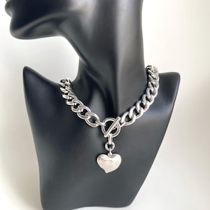 K&ouml;nnte beinhalten: Eine silberne Kettenkette mit einem gro&szlig;en, klobigen Gliederdesign. Die Kette hat einen Toggle-Verschluss und einen baumelnden Herz-Anh&auml;nger.
