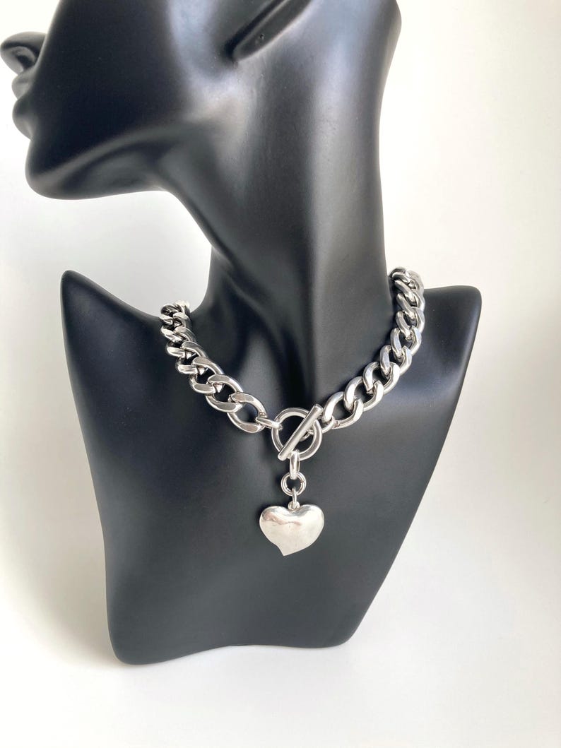 K&ouml;nnte beinhalten: Eine silberne Kettenkette mit einem gro&szlig;en, klobigen Gliederdesign. Die Kette hat einen Toggle-Verschluss und einen baumelnden Herz-Anh&auml;nger.