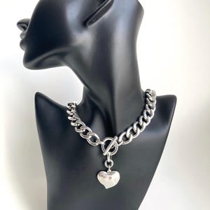K&ouml;nnte beinhalten: Eine silberne Kettenkette mit einem gro&szlig;en Herz-Anh&auml;nger. Die Kette ist dick und hat ein klobiger Kettenglied-Design. Der Herz-Anh&auml;nger ist glatt und hat eine leicht abgerundete Form.