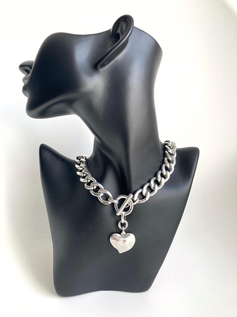 K&ouml;nnte beinhalten: Eine silberne Kettenkette mit einem gro&szlig;en Herz-Anh&auml;nger. Die Kette ist dick und hat ein klobiger Kettenglied-Design. Der Herz-Anh&auml;nger ist glatt und hat eine leicht abgerundete Form.