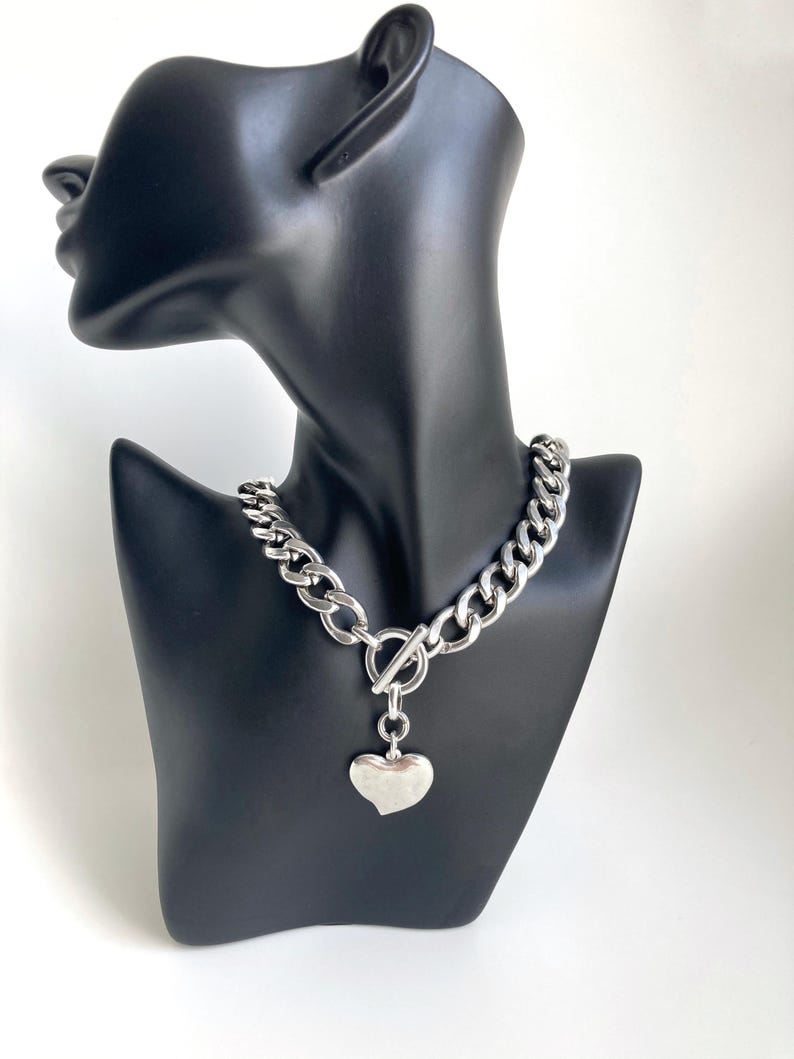 K&ouml;nnte beinhalten: Eine silberne Kettenkette mit einem gro&szlig;en, klobigen Gliederdesign. Die Kette hat einen Toggle-Verschluss und einen gro&szlig;en, silbernen Herzanh&auml;nger.