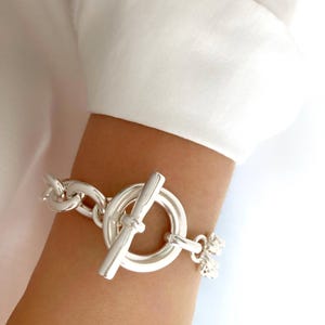 May include: A silver chain bracelet with a large, round toggle clasp. The bracelet is made of thick, chunky links.