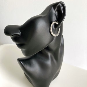 Peut inclure: Boucles d'oreilles créoles en argent avec une texture martelée. Les boucles d'oreilles sont présentées sur une tête de mannequin noire.