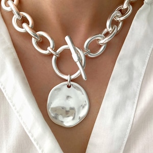 Puede incluir: Un collar de cadena gruesa de plata con un gran colgante redondo de plata martillado. El collar tiene un cierre de palanca.