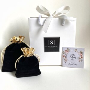 Puede incluir: Una bolsa de regalo blanca con una cinta de sat&eacute;n blanca y un lazo. La bolsa tiene un logotipo negro con la letra "S" y el texto "Silverhoopjewelry Accessories". La bolsa est&aacute; sobre una superficie blanca con dos bolsitas de terciopelo negro con cierres de cord&oacute;n dorado. Una tarjeta con el texto "Feliz cumplea&ntilde;os para ti! Contigo todo lo mejor" y un dise&ntilde;o de globos de colores tambi&eacute;n est&aacute; sobre la superficie.