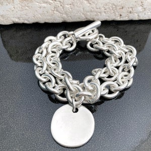 Peut inclure: Un bracelet argenté avec un design de chaîne à deux brins. Le bracelet est doté d'un fermoir à bascule et d'un charm rond et vierge. Le bijou est présenté sur une surface sombre.