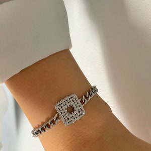 Puede incluir: Un brazalete de cadena de plata con un dije cuadrado con un dise&ntilde;o geom&eacute;trico.