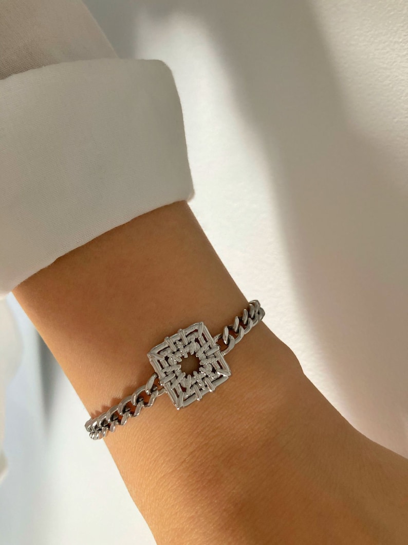Puede incluir: Un brazalete de cadena de plata con un dije cuadrado con un dise&ntilde;o geom&eacute;trico.