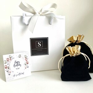Puede incluir: Una bolsa de regalo blanca con una cinta de sat&eacute;n blanca y un logotipo en blanco y negro que dice "Silverhope Jewellery Handmade". La bolsa est&aacute; sobre una superficie blanca con una bolsa de terciopelo negro con un cord&oacute;n de ajuste dorado y una tarjeta que dice "Feliz cumplea&ntilde;os para ti! Te deseo lo mejor".