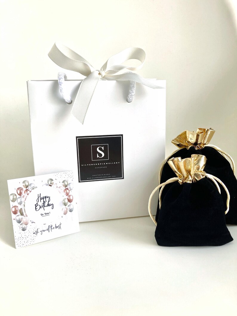 Puede incluir: Una bolsa de regalo blanca con una cinta de sat&eacute;n blanca y un logotipo en blanco y negro que dice "Silverhope Jewellery Handmade". La bolsa est&aacute; sobre una superficie blanca con una bolsa de terciopelo negro con un cord&oacute;n de ajuste dorado y una tarjeta que dice "Feliz cumplea&ntilde;os para ti! Te deseo lo mejor".
