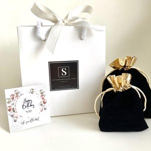 Puede incluir: Una bolsa de regalo blanca con una cinta de sat&eacute;n blanca y un logotipo negro que dice "Silver Shop Jewellery Handmade". La bolsa est&aacute; llena de una bolsa de terciopelo negro con un cord&oacute;n de ajuste dorado. La bolsa est&aacute; sobre una superficie blanca. Una tarjeta con el texto "Happy Birthday To You! Wish you off the best" tambi&eacute;n est&aacute; sobre la superficie.