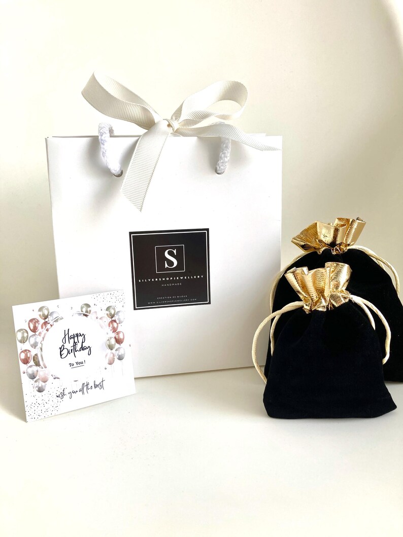 Puede incluir: Una bolsa de regalo blanca con una cinta de sat&eacute;n blanca y un logotipo negro que dice "Silver Shop Jewellery Handmade". La bolsa est&aacute; llena de una bolsa de terciopelo negro con un cord&oacute;n de ajuste dorado. La bolsa est&aacute; sobre una superficie blanca. Una tarjeta con el texto "Happy Birthday To You! Wish you off the best" tambi&eacute;n est&aacute; sobre la superficie.