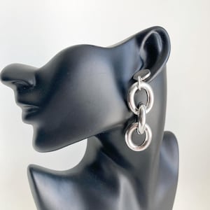 Peut inclure: Boucles d'oreilles pendantes argentées composées de trois anneaux circulaires reliés. Les boucles d'oreilles sont présentées sur une tête de mannequin noire, sur fond blanc. Finition polie et réfléchissante.