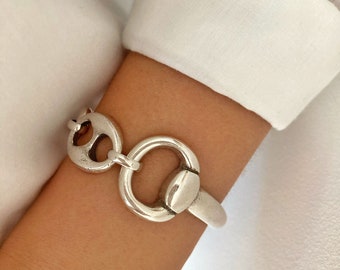 May include: A silver bracelet with a distinctive design. It features a large circular link, a smaller decorative piece, and a clasp. The bracelet is worn on a wrist, set against a white shirt.