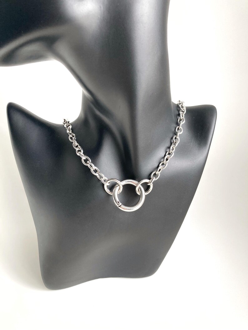 May include: A silver chain necklace with a large, round, silver pendant. The necklace is on a black mannequin.