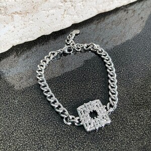 Puede incluir: Un brazalete de cadena de plata con un colgante cuadrado con un dise&ntilde;o geom&eacute;trico.