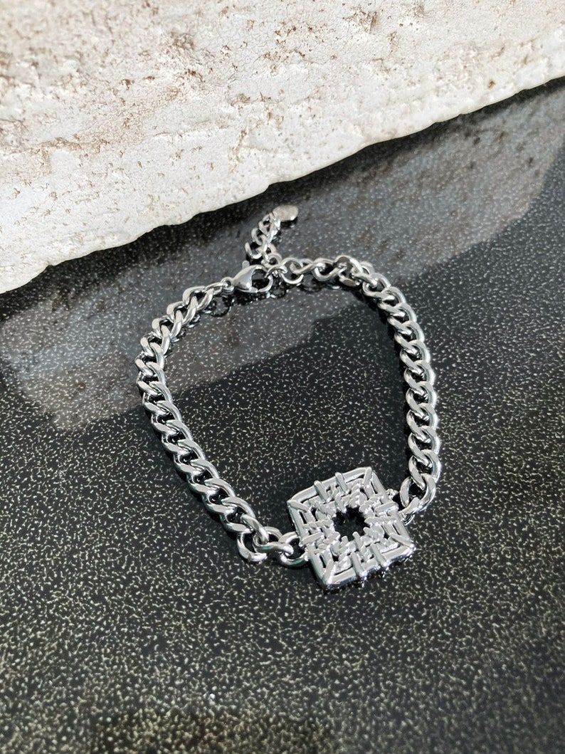 Puede incluir: Un brazalete de cadena de plata con un colgante cuadrado con un dise&ntilde;o geom&eacute;trico.