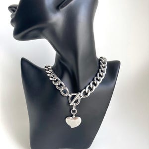 K&ouml;nnte beinhalten: Eine silberne Kettenkette mit einem gro&szlig;en, klobigen Gliederdesign. Die Kette hat einen Toggle-Verschluss und einen gro&szlig;en, silbernen Herzanh&auml;nger.