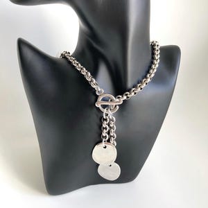 May include: A silver chain necklace with a toggle clasp and two dangling silver discs.