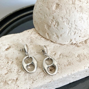 Könnte beinhalten: Ein Paar silberfarbene Ohrhänger. Jeder Ohrring hat einen bohnenförmigen Anhänger, der an einem Stecker hängt. Die Ohrringe werden auf einer strukturierten, cremefarbenen Steinoberfläche präsentiert.