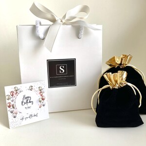 Puede incluir: Una bolsa de regalo blanca con una cinta dorada y un logotipo en blanco y negro que dice "Silver Shop Jewellery Handmade". La bolsa est&aacute; sobre una superficie blanca junto a una bolsa de terciopelo negro con una cinta dorada. Una tarjeta con el texto "Happy Birthday To You! Wish you off the best" est&aacute; en primer plano.