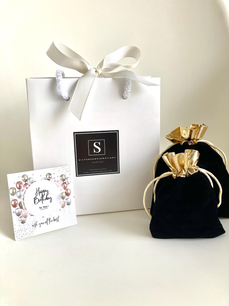 Puede incluir: Una bolsa de regalo blanca con una cinta dorada y un logotipo en blanco y negro que dice "Silver Shop Jewellery Handmade". La bolsa est&aacute; sobre una superficie blanca junto a una bolsa de terciopelo negro con una cinta dorada. Una tarjeta con el texto "Happy Birthday To You! Wish you off the best" est&aacute; en primer plano.