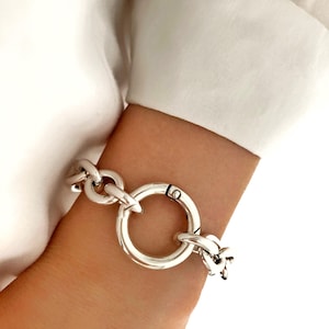 Op de afbeelding: Een zilverkleurige armband met een grote ronde sluiting en dikke schakels. De armband wordt om een pols gedragen, met een witte mouw op de achtergrond. Het ontwerp van de armband is opvallend en modern.