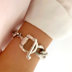 Peut inclure: Un bracelet argenté avec un fermoir à bascule. Le bracelet présente un design de chaîne épaisse avec un fermoir rectangulaire et une fermeture à barre incurvée. Le bracelet est porté sur un poignet, contre une chemise blanche.