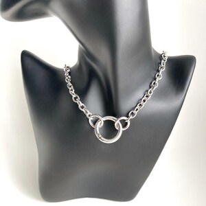 May include: A silver chain necklace with a large, round, silver pendant. The necklace is on a black mannequin.