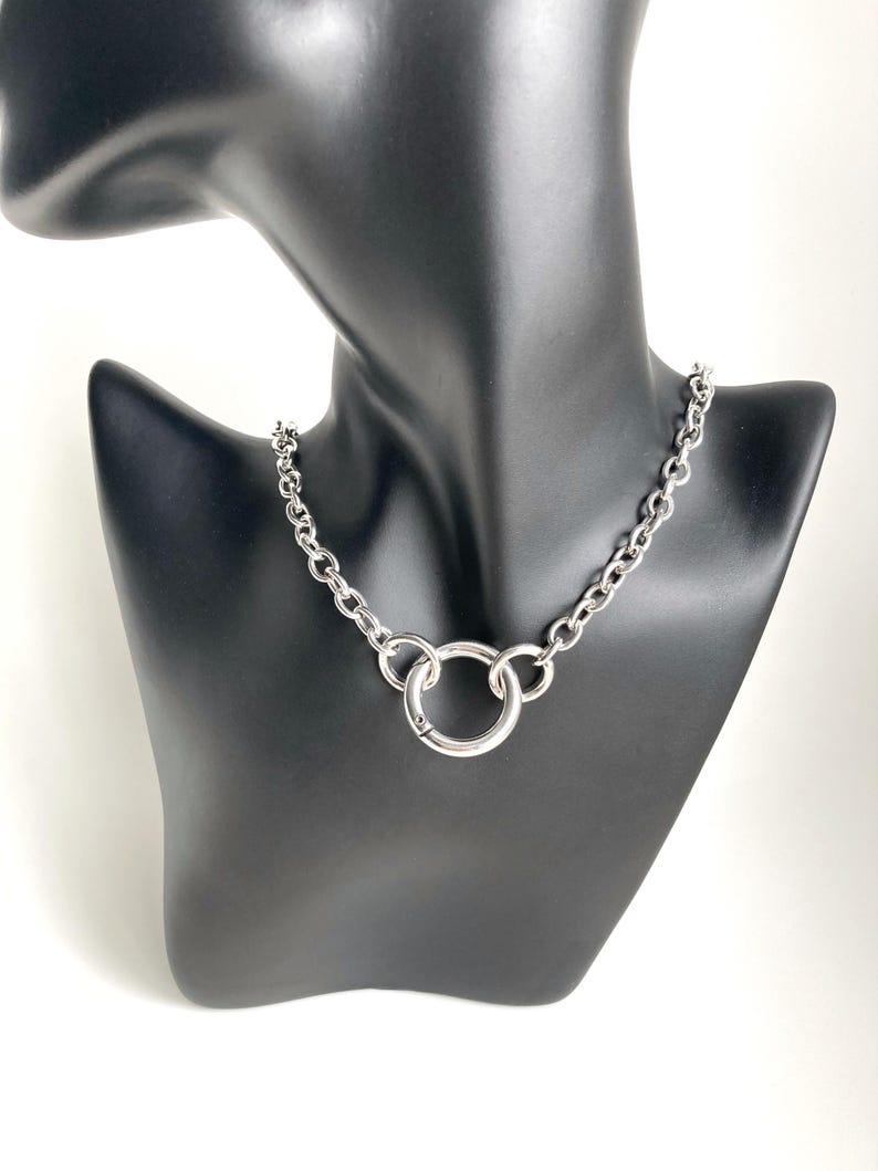 May include: A silver chain necklace with a large, round, silver pendant. The necklace is on a black mannequin.