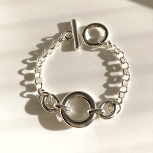 Op de afbeelding: Een zilveren armband met een tuimelsluiting. De armband heeft een kettingontwerp met een groot cirkelvormig middengedeelte en een kleinere ring aan elke kant. De tuimelsluiting is een staaf die in een cirkelvormige lus past.