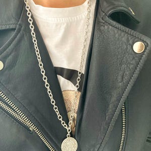 Puede incluir: Un collar de cadena de plata con un gran colgante redondo de plata. El colgante tiene un diseño detallado y cuelga de una cadena de plata.