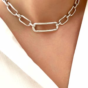 Peut inclure: Un collier en chaîne argentée avec un grand pendentif rectangulaire. La chaîne est faite de métal épais et lié.